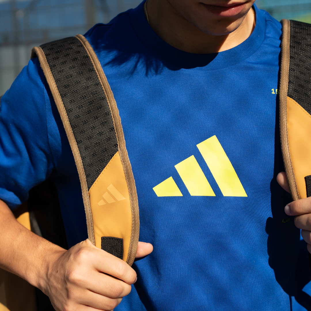 Person wearing a blue Adidas shirt with a yellow logo, holding a backpack.